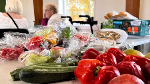 A table with a mix of fruit and vegetables on it, including red peppers, cucumbers and courgettes.