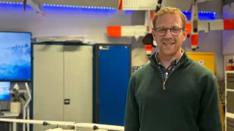 Toby King-Cline has short ginger hair and stubble, and is wearing rounded glasses. He has a blue shirt on that is mostly covered by a green quarter-zip. Out of focus behind him is a workshop.