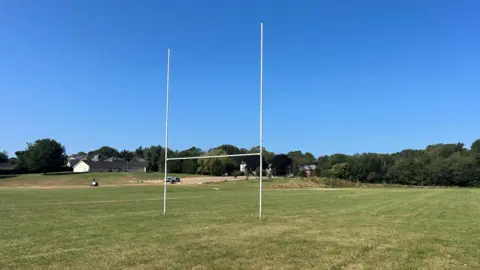 A low down image of rugby posts looking up at them on a clear blue day. The pitch is visible as well as trees in the background.