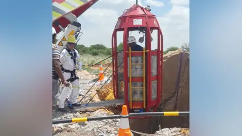 Giwusa A man in a white kettle stands next to a crane about to lower a cage down a mine shaft.