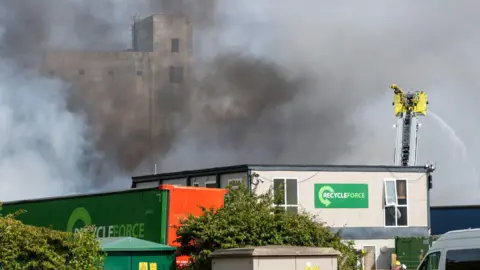 Fire crews responding to a blaze at a recycling waste processing facility on an industrial estate in Corby. Black smoke is coming off a building and billowing into the air. A firefighter is on an aerial ladder.