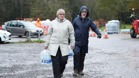 PA Media Two people carrying bottles of water through a muddy car park