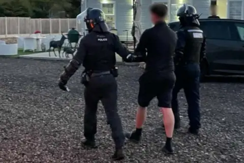 Essex Police Shot from behind of one of the gang members being led away, with a police officer at either side. The back of his head is blurred out and he is wearing a black fleece, shorts, ankle socks and slider. The officers wear protective raid uniform. They walk along a pebbled path with a caravan and car partially seen in the background.