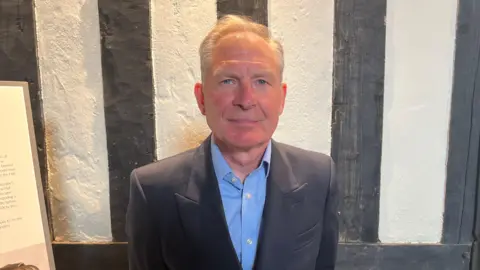 A man stands in front of a white wall with black beams. He is wearing a navy blazer and a blue shirt. He has short, grey hair and is looking straight into the camera.