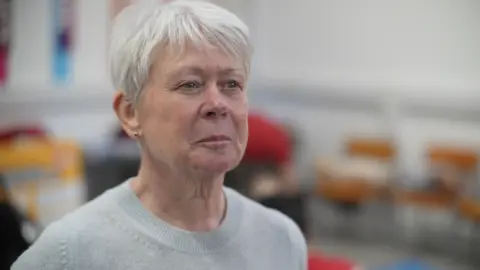 Jamie Niblock/BBC Carol Wood is looking to the right of the camera. She has short grey hair and is wearing a grey long-sleeved jumper.