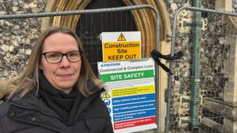 Andrew Turner/BBC Helen Orford stands outside the entrance of St John's Church, where signage warns of the dangers inside. There is temporary fencing around the building. Ms Orford is wearing a black scarf and a black coat, with a faux fur hood lining. She is wearing black rimmed glasses and has long blonde hair.