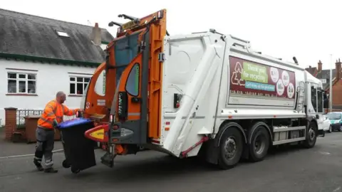 Getty Images A man loading a recycling bin with a blue lid into the back of a bin lorry.