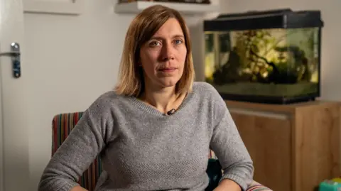 BBC Catriona Ball sits on a striped armchair. There is a brown cupboard and fish tank behind her. She is wearing a grey jumper. She has brown/blonde hair in a bob.