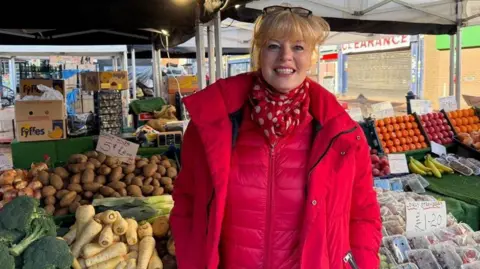 BBC Woman wearing a bright pink coat and scarf smiling at the camera, stood in front of a fruit and veg stand.
