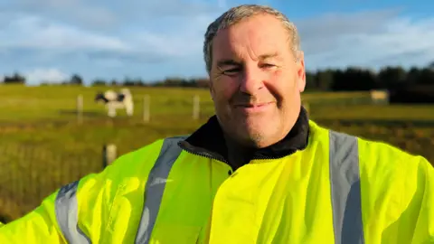 BBC Mike Duxbury smiling at camera, he is wearing a bright yellow safety jacket, there is a cow in a field in the distance behind him.