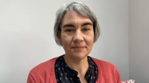 Elliot Deady/BBC Margaret Winchcomb looks directly at the camera with a neutral expression. She is wearing a dark pink cardigan over a blue patterned blouse. She has short grey hair and is sitting in front of a white or grey wall.