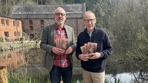 Two men are holding brown paper books while standing up looking at the camera. Behind them are buildings and a lake.