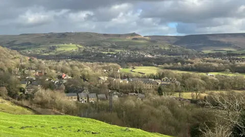 A green bank gives way to a vista of rolling hills and moorland, dotted with trees, with a small spires and the rooftops of homes seen in amongst the trees.