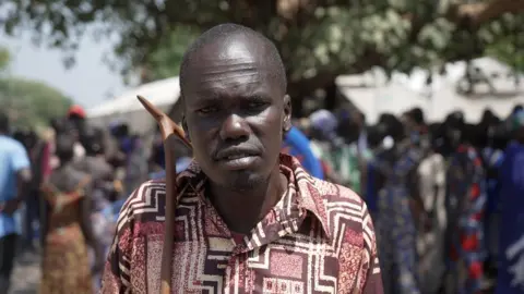 Hoth Wan Kornyom looks at the camera. He has a maroon and cream patterned shirt and is holding a wooden stick.