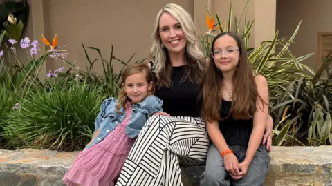 Keri Bird Keri Bird, a blonde woman wearing a black top and geometric patterned trousers, is sitting between her two children. On the left, her daughter with brown hair is wearing a pink dress and denim jacket, cuddled into her mum. On the right, a slightly older girl with brown hair and glasses is wearing a black vest top and grey trousers. 