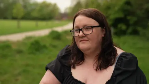 A woman with shoulder length brown hair and black glasses wearing a black short sleeved top stands on a path in a green area