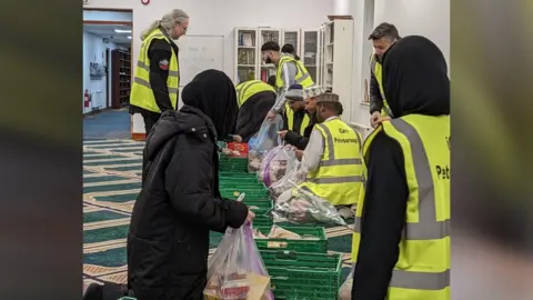 Volunteers wearing green high-visibility jackets- packing food parcels at a room inside the Mosque