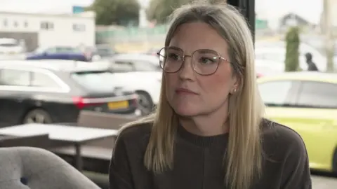 A woman looks slightly to the side, she does not smile. She has blonde hair and wears glasses, and a brown knit jumper. She is sitting inside a café and there's a big window behind her. There is a car park in the background.