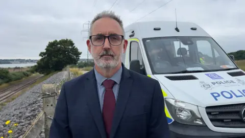 John Fairhall/BBC Det Supt Mike Brown in a blue suit and burgundy tie, standing beside a railway track and with a police van behind him.