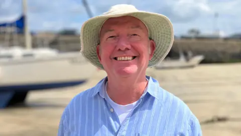 Steven Mortimer, an older man, is wearing a light-coloured bucket hat, plus a blue and white striped shirt over a white T-shirt. He is smiling at the camera.