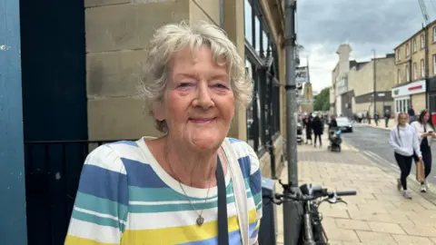A woman with short white curly hair smiles towards the camera, wearing a yellow, white and blue striped t-shirt. She is also wearing a necklace and has two bag straps over her shoulder. She is stood on a pavement where there are people walking in the background.