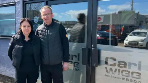 Vikki Irwin/BBC Carl and April Wigg are standing outside their workshop. There is a glass double door with Carl Wigg Auto Services written in the glass. Cars are reflected in the glass. Both Carl and April are dressed in black, wearing black gilets.
