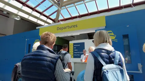 Passengers queuing to go through departures at Jersey's ferry terminal. The photo is taken over the shoulder of the passengers.