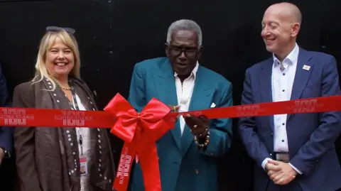 A line of two men and one woman stand behind a red ribbon, which has a large bow in the middle. Rudolph Walker is in the middle, cutting the ribbon. He wears white trousers an shirt and a teal jacket. The room they are in is dark.