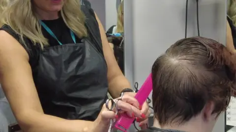 A person wearing a black apron and holding a pink comb to someone else's hair. They are holding scissors and seem to be about to cut the person's hair.