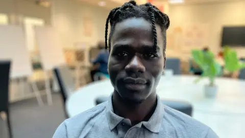 Salah from Sudan, who is wearing a grey polo shirt, stands in a conference room in front of a large circular table with a potted plant in the centre.