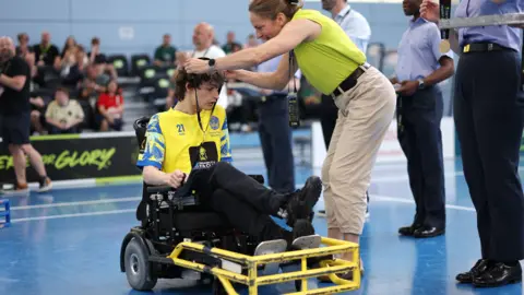 The FA via Getty Images Dan gets his winners medal placed around his neck on court. He wears a yellow shirt. He has dark hair and is sat in his wheelchair. Several fans are pictured, sat, in the background.