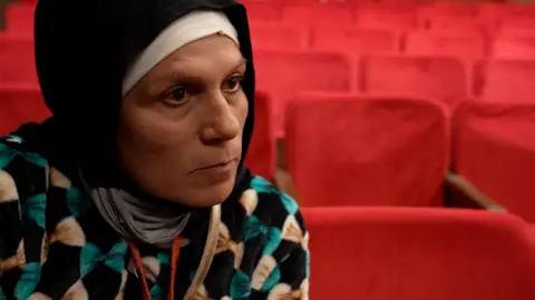 A woman wearing a hijab stares with a blank expression off camera, wearing a jumper with a pattern of bows on it, inside a theatre with red seats.