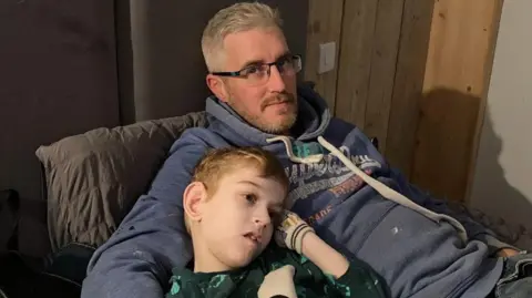 Band of Builders Shaun and Toby slouched on a sofa together, with the dad's right arm around his boy. Shaun has short grey hair and stubble and is wearing glasses and a blue Superdry hoodie. Toby has short blond hair and is wearing a green top and beige coloured gloves.