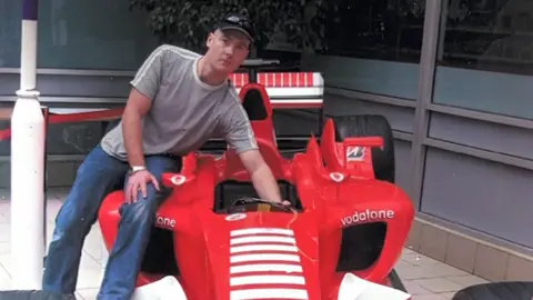 Lucy Early A man is on a racing car that is mainly red on the photo. He is wearing a black cap, a grey T-shirt and blue jeans.