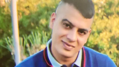 West Yorkshire Police Choudry sitting on an outdoor bench, wearing a short‑sleeved navy blue polo shirt with red and white trim on the collar and sleeves. The shirt has a circular white emblem on the left side of the chest. He is also wearing blue jeans and a wristband on their right wrist.