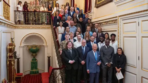 Aaron Chown/PA Media Martin and Tara Cosser (4th row, 2nd and 3rd from left) standing on steps of a staircase at St James's Palace with the King, Prime Minister and Idris Elba at the front