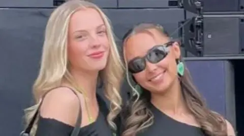 A woman with long blonde hair stands beside another with tied up dark hair both are smiling toward the camera