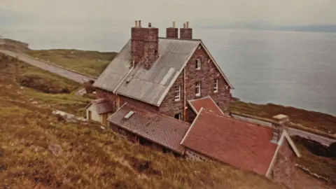 Hostelling Scotland A large sandstone brick building with a tall chimneys on its steeply peaked roof. The hotel overlooks a large loch.