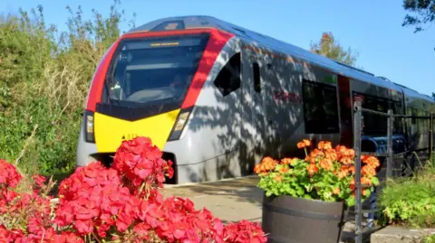 A Greater Anglia train, which is grey with red and yellow stripes at its front, is drawn up alongside a station platform. There are red and orange flowers in the foreground. It is a bright, sunny day and the sky is blue. 