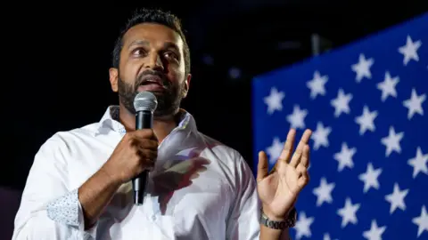 Getty Images Kash Patel, who has short brown hair and a beard, and is wearing a white dress shirt with the collar unbuttoned and sleeves slightly folded, gestures with one hand as he speaks into a microphone held in his other hand