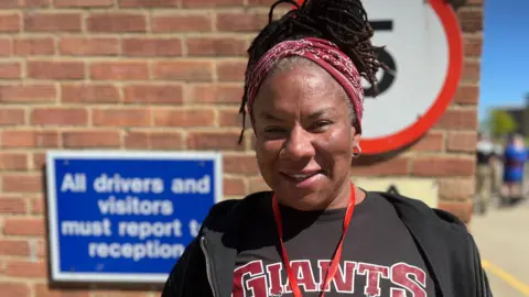 Lauren Carter/BBC Andrea Thompson, wearing a black T-shirt with the word GIANTS printed on it in red, and wearing a black hoodie. She wears a red headscarf or bandana. There is a brick wall behind her. She is outside on a sunny day.