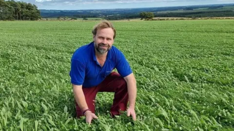 Simon Bainbridge Simon Bainbridge is a man in his 40s with a beard and is wearing a blue top and reddish trousers. He is kneeling down in a field of crops and behind him several miles of fields and woodlands are visible 