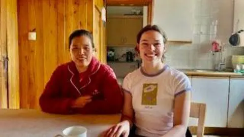 CamSight Lucy and Jean are sitting together at a kitchen table and are both smiling at the camera. Jean has her arms crossed and is wearing a red long-sleeved top. It has a collar which has been lined around the edges in white. She has black hair that has been tied back. Lucy has her hands resting on her knees. She is wearing a white top with a pink line around the sleeves and collar. It also has a green rectangle in the centre.