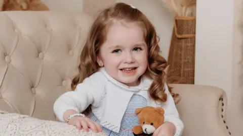 Joanne Watters Photography Hollie pictured on a cream chair, the photo is professional. She has long curly red/brown hair and is wearing a blue and white dress with a white cardigan. She is also holding a teddy bear. 