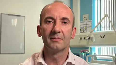 A man with a pink shirt in a hospital ward with a whiteboard and a bed behind him and a window with blinds