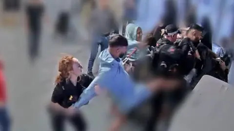 A man in blue kicks in the direction of a police officers while a police officer with red hair holds his arms behind him at the car park pay stations at Manchester Airport.