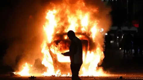 A fire burns in the front of the photo, to the right a line of police in riot gear can be seen and behind them in the distance a crowd of people is visibile