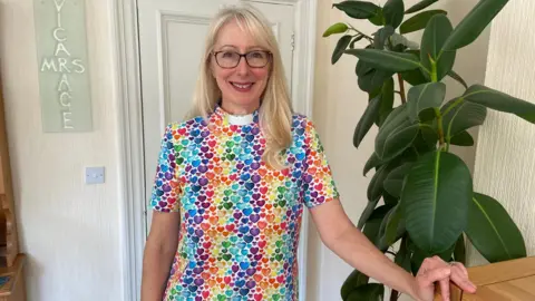 Tanya Lord A woman with long blonde hair and glasses smiles as she stands in a room with beige wallpaper next to a cheese plant. She is wearing a short-sleeved top with rainbow-coloured hearts and a clerical white collar.