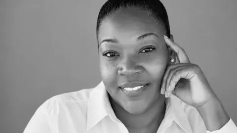 A black and white photo of a woman in a white blouse smiling into the camera. She has her hair drawn back off her face.