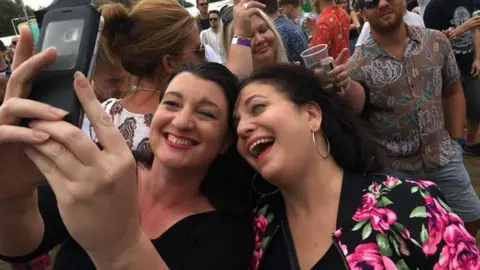 The image shows two women taking a selfie together. They are in a crowd with other people. Both have dark hair and are smiling. The woman on the left, holding the phone, is wearing a black top. The woman on the right is wearing a black t-shirt and a black jacket with bright pink flowers on it